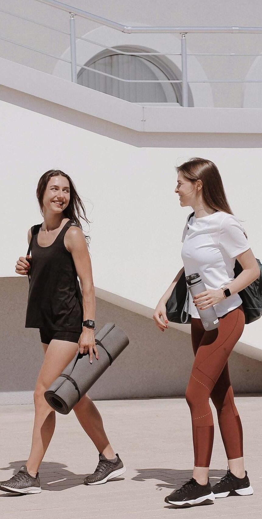 Two women in active wear walking outdoors while smiling and carrying yoga mats.