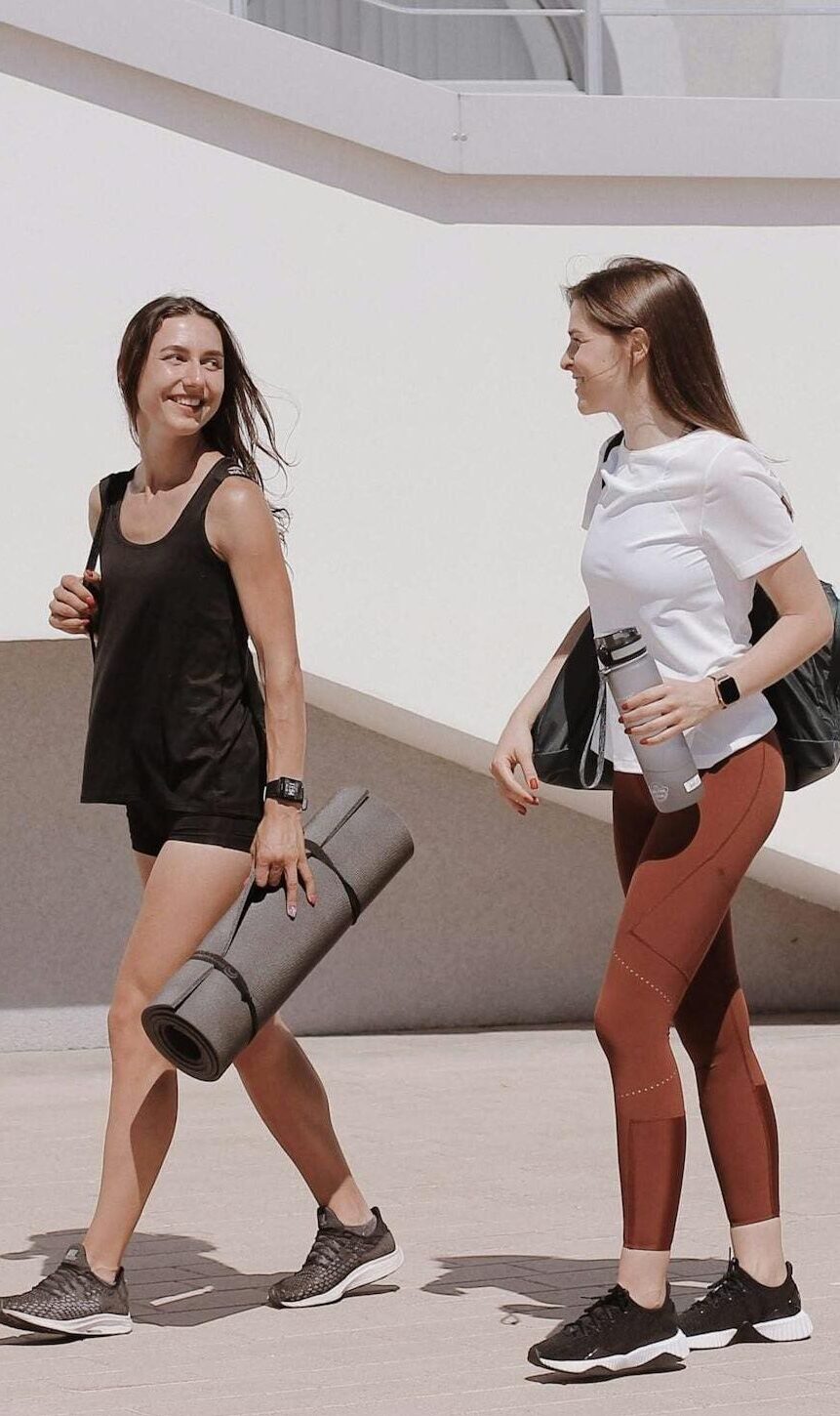 Two women in active wear walking outdoors while smiling and carrying yoga mats.