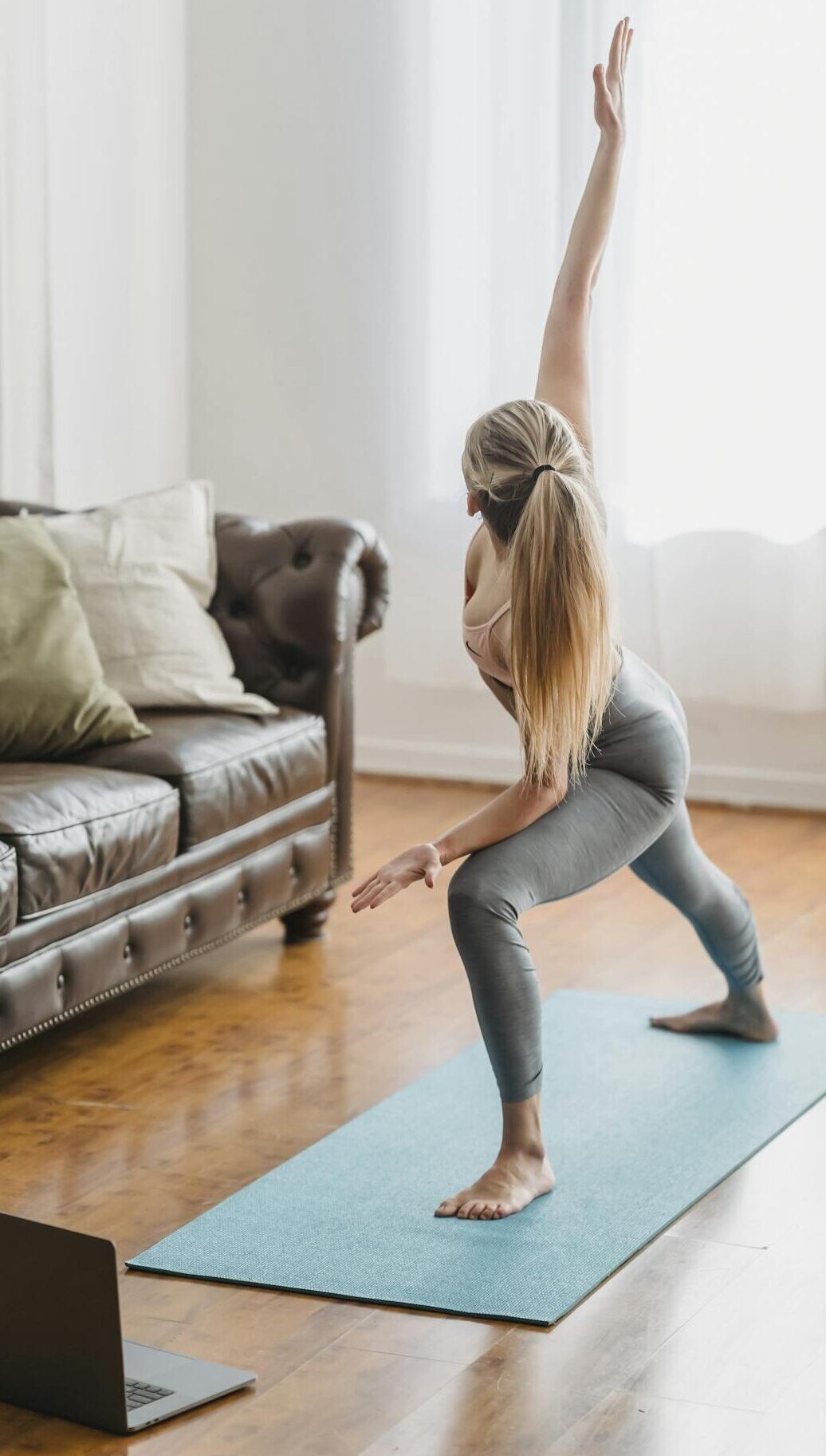 Full body side view of unrecognizable female in activewear practicing extended side angle posture while performing yoga exercise in living room with laptop